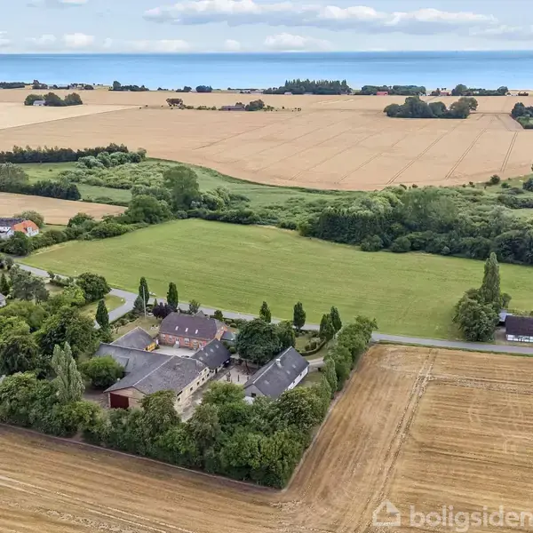 Et landligt landskab med en klynge bygninger omgivet af træer på en gård. Omkringliggende marker strækker sig mod horisonten med havet i baggrunden under en blå og skyet himmel.