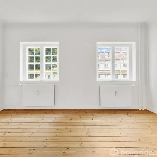 Et lyst rum med to vinduer, der lukker dagslys ind, og en trægulvsoverflade. Væggene er hvide, hvilket giver et rent og enkelt udseende.
