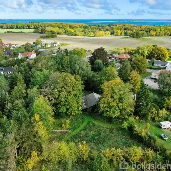 Huse blandt træer i landsby. Ovenfra synlig have med en enkelt campingvogn. Baggrund med marker og skov. Miljøet er fredeligt og idyllisk.