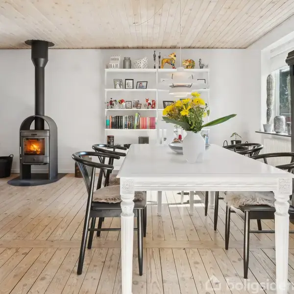Et spisebord med blomsterbuketten står centralt placeret i en lys stue med trægulv. Der er en brændeovn i hjørnet og en sort skab ved væggen.