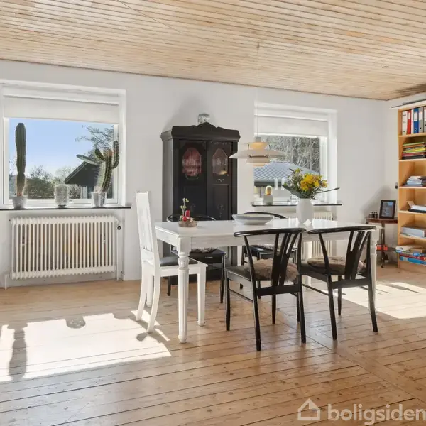 Et hvidt spisebord med sorte stole står i en lys stue ved siden af en varm pejs. Vasen på bordet indeholder gule blomster. Reol, hvide vægge, trægulv.