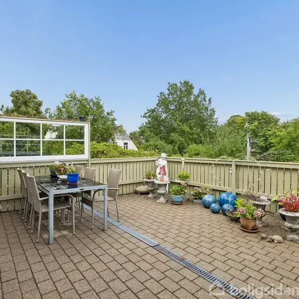 Terrasse med flisebelægning og havemøbler; omgivet af grønne planter og træer; dekoreret med krukker, blå kugler og en hvid statue.