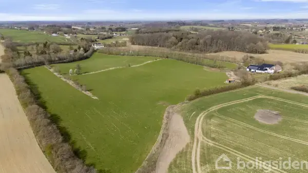 En stor, grøn mark ligger ubenyttet omgivet af træer og marker. I baggrunden ses flere huse og en skov i det fjerne.