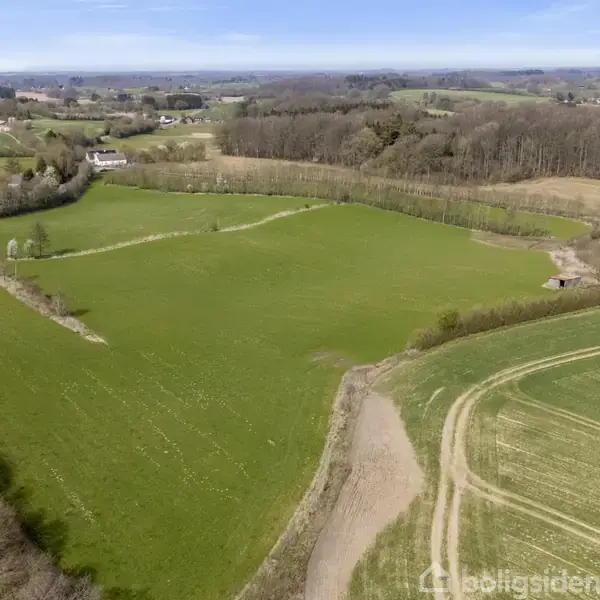 En stor grøn mark omgivet af skove og marker. En hvid bygning ligger i højre side. Landskabet strækker sig ud i det fjerne under en klar himmel.