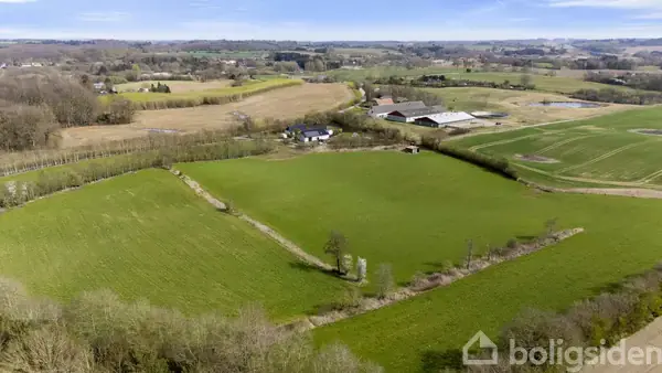 Grønne marker strækker sig over et bakket landskab. En lille landejendom med flere bygninger ses i midten, omgivet af skove og marker under en klar blå himmel.