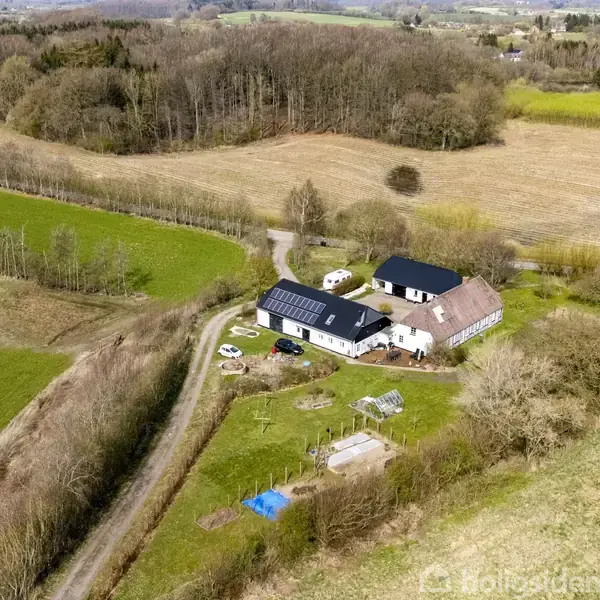 Luftfoto af en gård omkranset af marker og træer. Bygningerne har solceller på taget. En grusvej fører til gården, omgivet af grønt landskab.