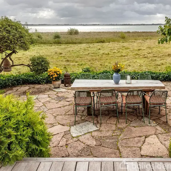 Et betonbord med otte stole står på en stenterrasse i haven med udsigt over en sø. Der er planter rundt om terrassen, og himlen er overskyet.