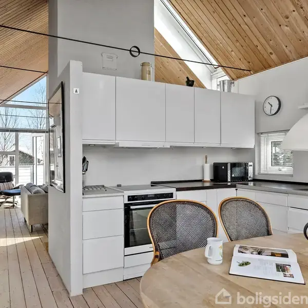 Moderne køkken med hvidt skabsværk og sort bordplade. Rundt bord med stole foran, kande og magasin ovenpå. Lysindfald fra skrå vinduer og hyggelig stue i baggrunden.