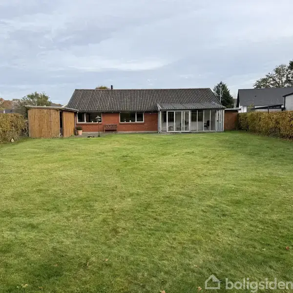 Et rødt murstenshus med sort tag. Flere store vinduer vender mod en velplejet græsplæne omgivet af hække. I højre side er en overdækket terrasse.