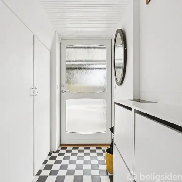 En entré med hvid skabsdør og skuffer; sort-hvidternet gulv. En oval spejl hænger på væggen ved siden af en yderdør med frostet glas. Ingen tekst fundet.