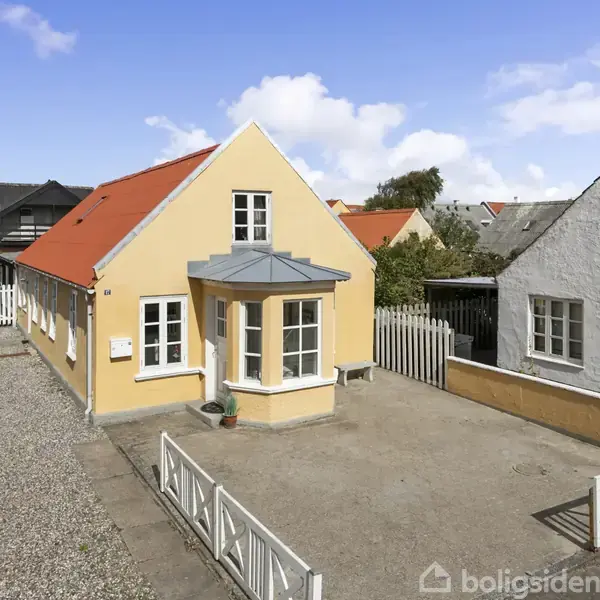 Et gult, vinkelbygget hus med rødt tag og blå frontudhæng står på en grusbelagt grund. Omgivet af hegn, ses det i en rækkehusområde med flere lignende bygninger.