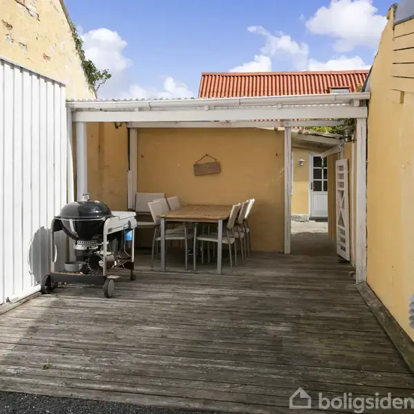 Grill står på terrasse under halvtag omgivet af gule vægge. Ved siden af står et spisebord med stole. Himlen er blå med skyer.
