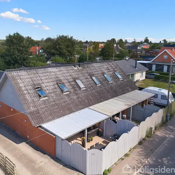 Stue med sort sofa og hvide borde dekoreret med rosa lys, hvor spisebord med rosa stole står ved siden af køkkenø. Moderne lysekrone hænger over spisebordet, og væggen har hvid skænk og TV.