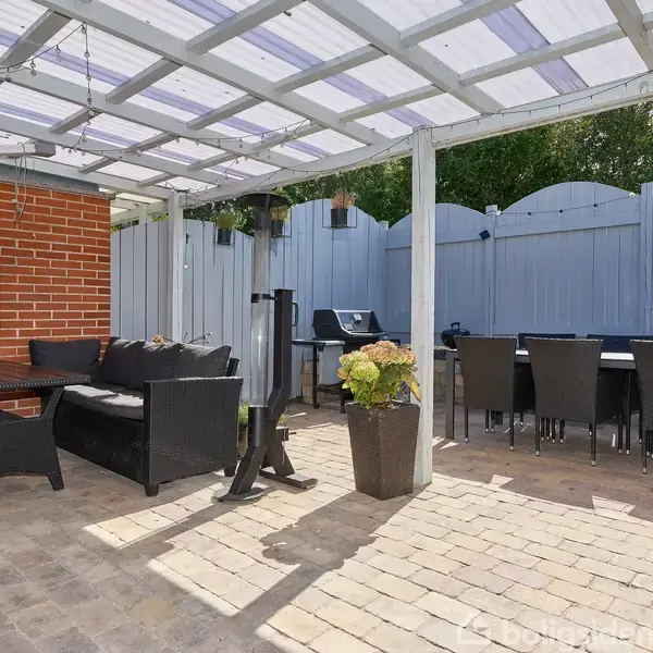 Terrasse med polyrattanmøbler og et bord, arrangeret under en overdækning ved siden af en murstensvæg. Grill og planter står på flisebelagt gulv, omkranset af et hvidt plankeværk.
