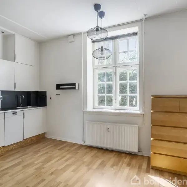 Køkken med hvide skabe og sort bordplade, placeret i et lyst rum med trægulv. En kommode står ved siden af et stort vindue med gardiner.