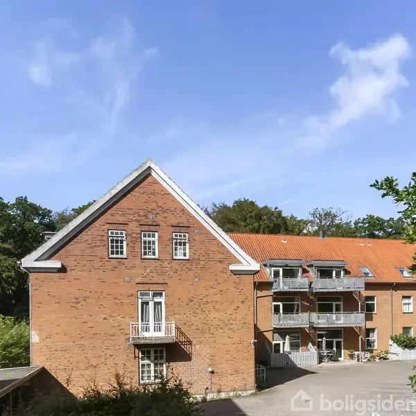 Rødstensbygning med flere balkoner, der strækker sig over to etager, omgivet af træer og parkeringsområde. Klar blå himmel i baggrunden.