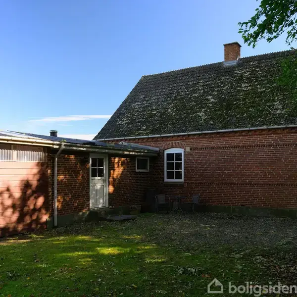 En rødstensbygning med et tilhørende skur ses i en grøn have. Huset er omgivet af skygger fra træer, og himlen er klar og blå.