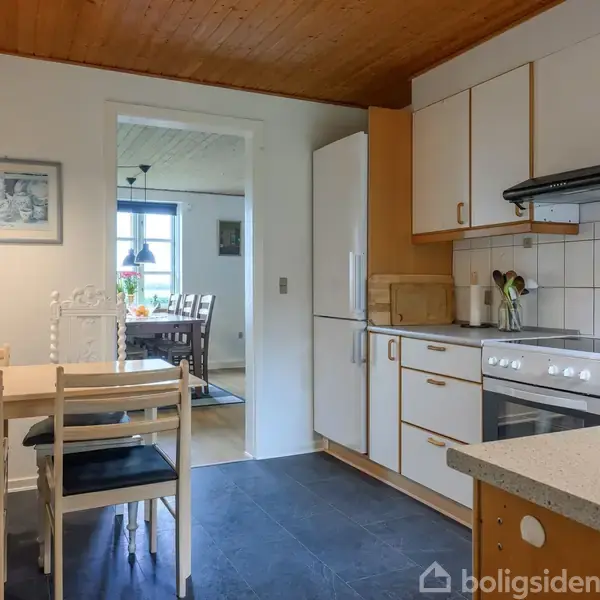 Et køkken med spisebord og stole i forgrunden; komfur og køleskab langs væggen. Døråbning fører til en spisestue med bord, stole og dekorationer.