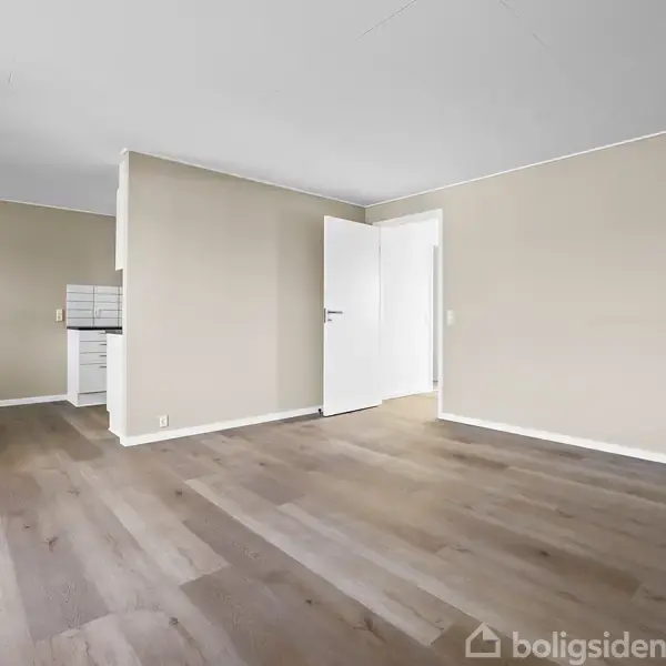 Et tomt rum med trægulv og beige vægge. En dør står åben, og til venstre kan et køkken skimtes. Væggene er lyse, og der er god plads.
