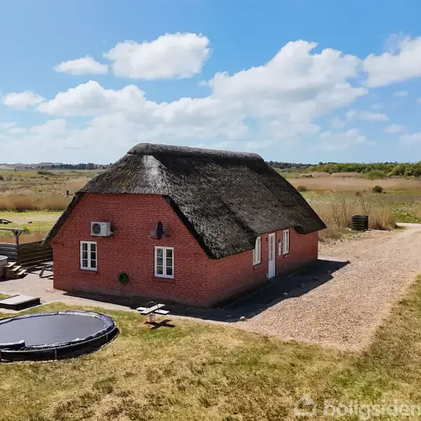 Rødt murstenshus med stråtag står på græsplæne. Nær ved ses en trampolin og et udendørs badekar. Landskabet omkring er åbent og fladt under en skyet himmel.