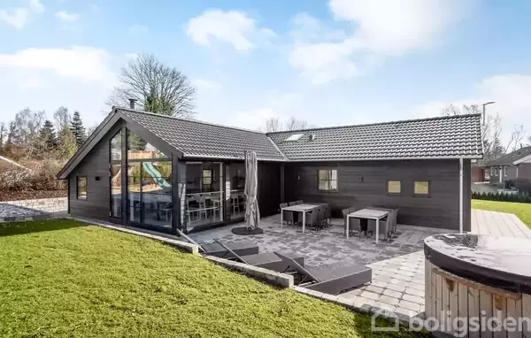 Et moderne sommerhus med store glaspartier står på en flisebelagt terrasse. Ved siden af er et bord og stole. Haven omkring huset er grøn og velplejet.