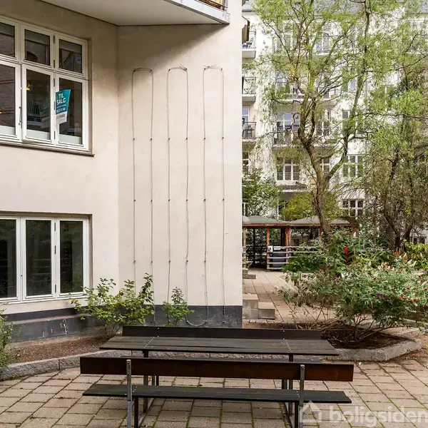 En udendørs bænk står på en flisebelagt terrasse foran en hvid bygning med store vinduer. Omkranset af buske og træer, skaber det en hyggelig gårdhaveatmosfære.