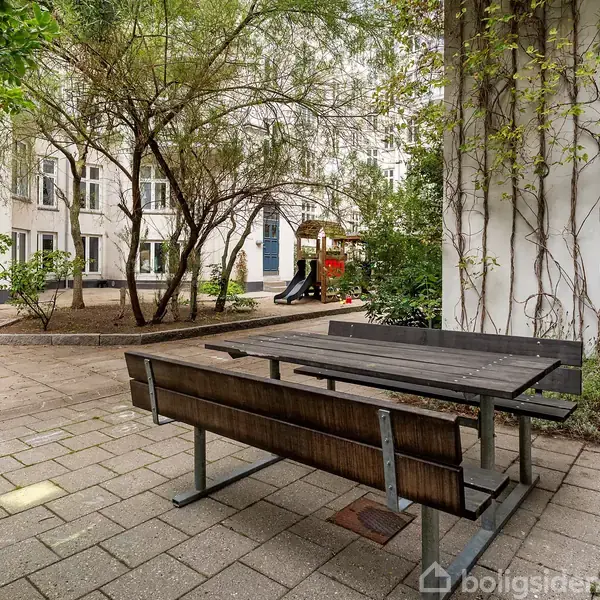 Et picnicbord står på en brostensbelagt gårdsplads omkranset af grønne planter. I baggrunden ses en legeplads med rutsjebane foran en bygning med store vinduer.