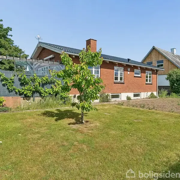 Rødt murstenshus med gavl og skorsten står stille omgivet af en velplejet have med grøn græsplæne, et lille træ og buske. Til venstre ses en pergola med planter.
