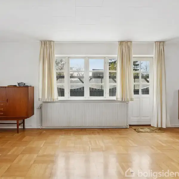 En stue med trægulv indeholder et vinduesparti med hvide gardiner og en dør. Møbler omfatter et spisebord, stole og skabe. Udenfor ses klare, blå himmel og træer.