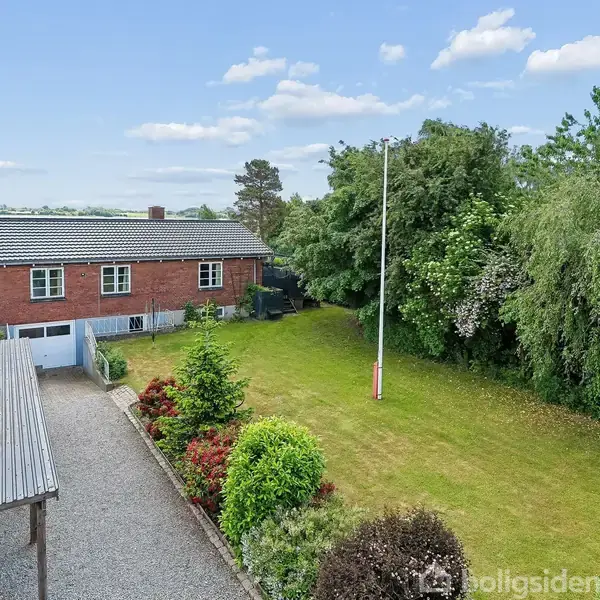 En rød murstenshus med kælder står stille i en have med grøn græsplæne og frodige buske. Til venstre ses en grusindkørsel med carport.