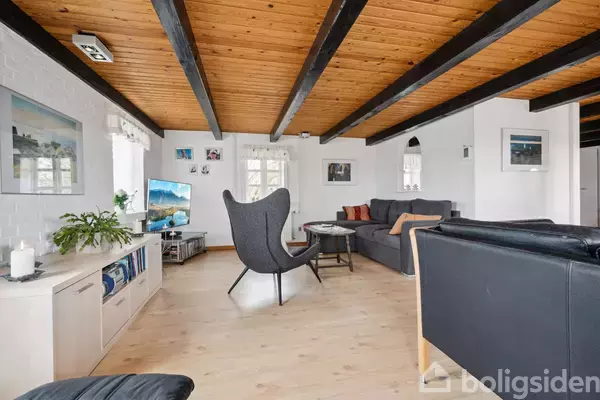 Stol står i stue med trægulv og loft. Rundt om er sofaer, et bord og et tv. Væggene har kunstværker, og der er naturligt lys fra vinduer.