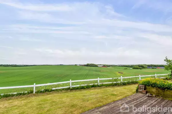 En grøn mark strækker sig ud i horisonten bag et hvidt stakit. Foran er en velplejet græsplæne og træterrasse. Himlen er let overskyet.