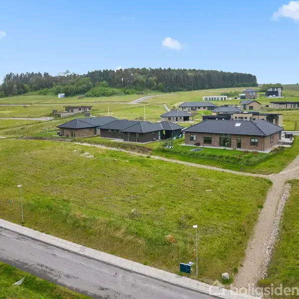 Huse står stille på grønne grunde i et nybygget boligområde omgivet af veje og lygtepæle, under en blå himmel med få skyer.