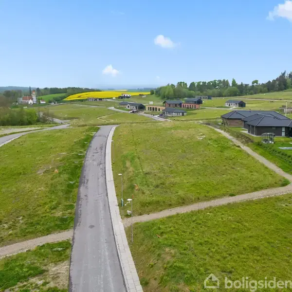 Vej strækker sig gennem grønt landskab, omkranset af åbne marker og enkelte moderne huse til højre. Skov og marker er synlige i baggrunden under en blå himmel med spredte skyer.