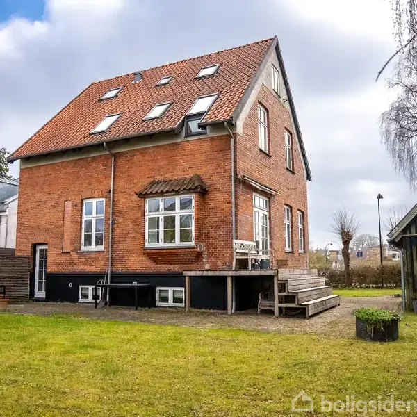 Rødt murstenshus med stejlt tag og mange vinduer, står på en velplejet græsplæne. Der er en trappe ved siden af huset, og en lille træbygning til højre. Skyet himmel.