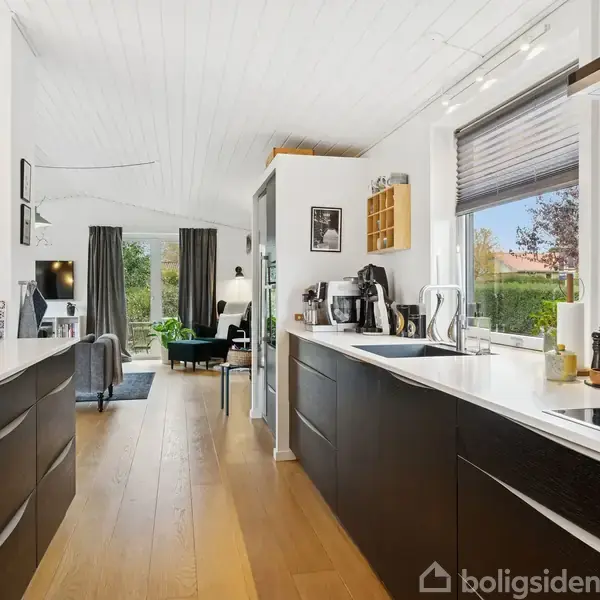 Et moderne køkken med mørke skabe og hvide bordplader, udstyret med kaffemaskine og komfur. I baggrunden er stue med sofa, tv, og store vinduer med udsigt til have.