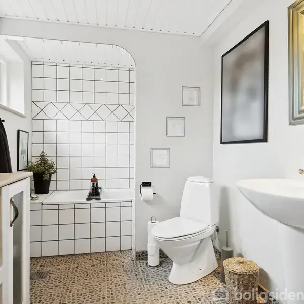 Badeværelse med klinkegulv og hvidt interiør; toilet ved siden af vask med spejl over. Badekar i baggrunden med dekorative fliser. Grønt plantepynt ved vinduet.