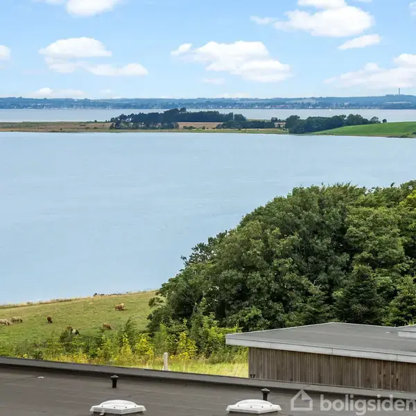 En tagterrasse med udsigt over en sø, grønne områder og en skov. I baggrunden er der flere små øer under en blå himmel med spredte hvide skyer.