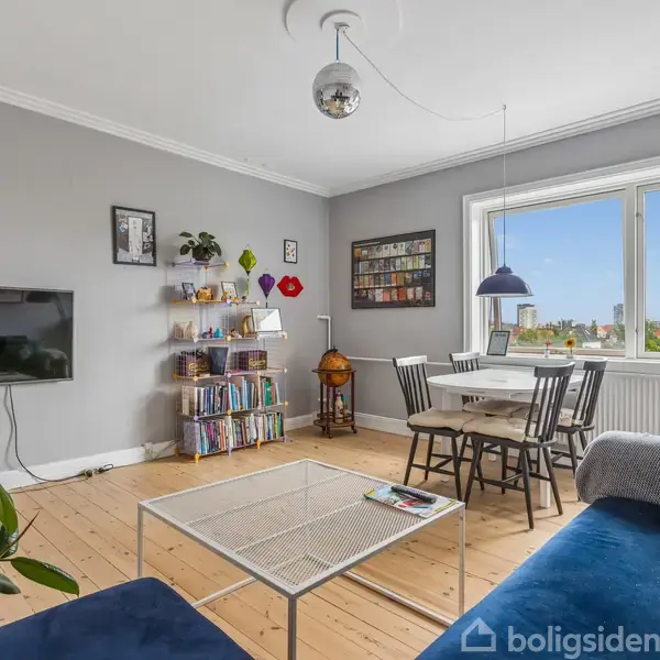 Stue med blå sofa og glasbord foran. Væghængt TV til venstre, bogreol i midten med flere bøger. Spisebord og stole ved vindue, grønne planter omkring.