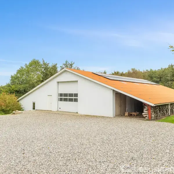 En stor hvid lade med orange tag står stille, omkranset af grønne træer og en grusbelagt forplads. Himmelen er blå og klar.