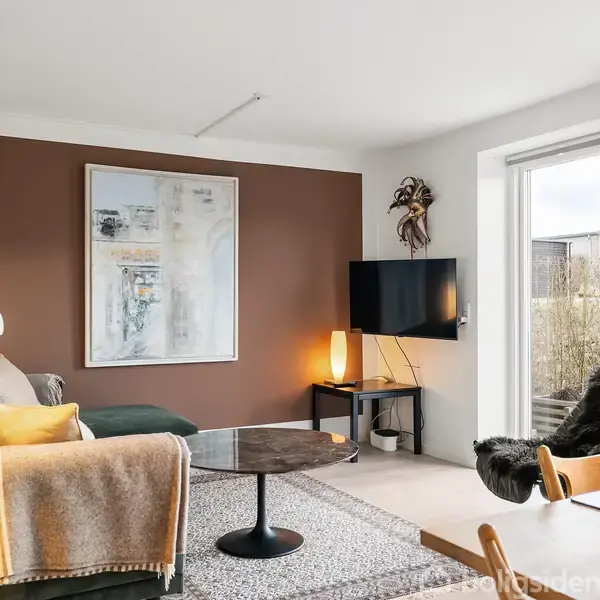 En stue med en grøn sofa placeret ved en brun væg dekoreret med kunst. Et rundt sofabord står foran sofaen. I hjørnet er et TV på væggen og en gulvlampe ved siden af. Vinduerne lader lys strømme ind, mens stole står ved spisepladsen.