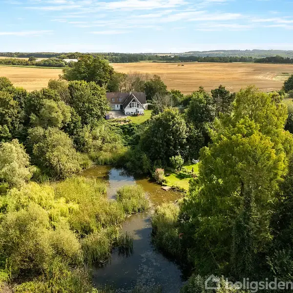 Et hus ligger omgivet af frodig skov og tæt ved en lille sø. I baggrunden ses åbne marker under en klar blå himmel.