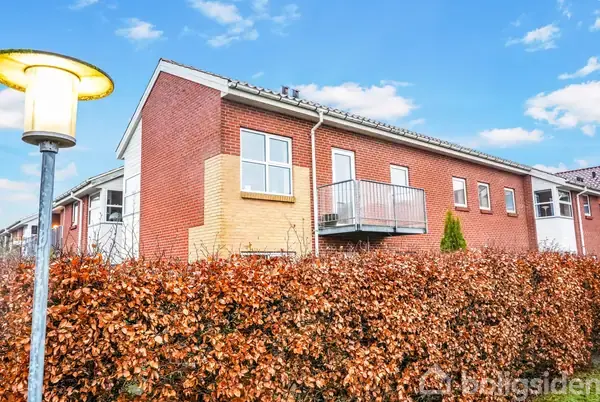 Et rødt murstenshus med en altan har en hæk foran. En gadelampe lyser i forgrunden under en blå, skyfri himmel.