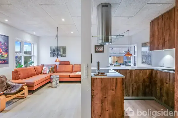 Orange sofa står i hjørnet af stuen. Rustik lænestol til venstre. Køkkenet har træskabe og emhætte. Store vinduer og kunst på væggene skaber et lyst og indbydende miljø.