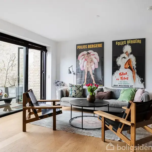 Stue med moderne møbler, herunder en sofa og to stole, står omkring et rundt sofabord. Store vinduer giver udsigt til en terrasse. Plakater med teksten "FOLIES BERGÈRE" hænger på væggen.