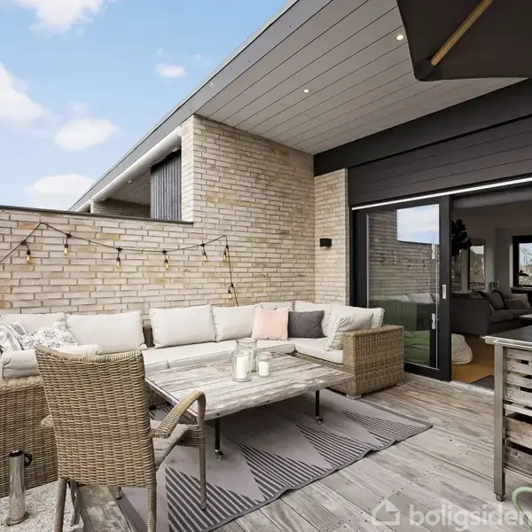 Terrasse med loungesæt og bord i træ omgivet af murstensvægge. Lyskæde hænger langs væggen. En dør åbner ind til en moderne stue. Himlen er klar med få skyer.