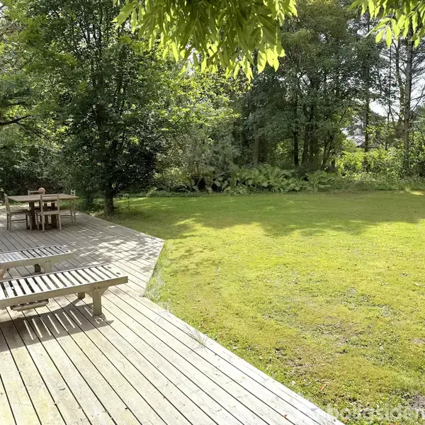 To liggestole står på en træterrasse i en have. I baggrunden er der en græsplæne omgivet af tætte træer, som skaber en skovlignende atmosfære.