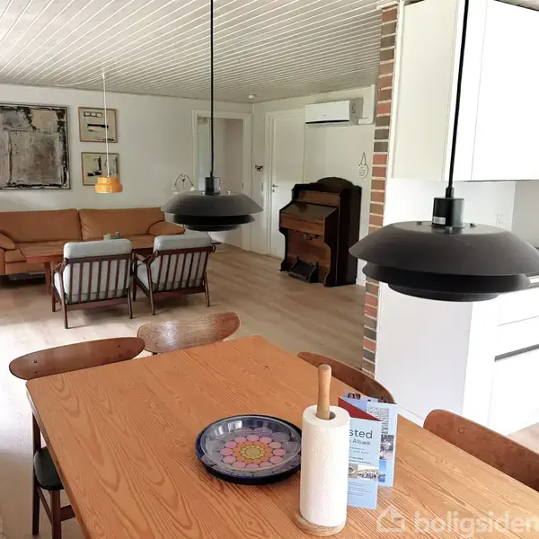 Spisebord med dekorativ tallerken og køkkenrulle, stående ved forgængerlamper. I baggrunden ses en stue med sofa, lænestole og et gammelt klaver. Væggene har indrammede billeder.
