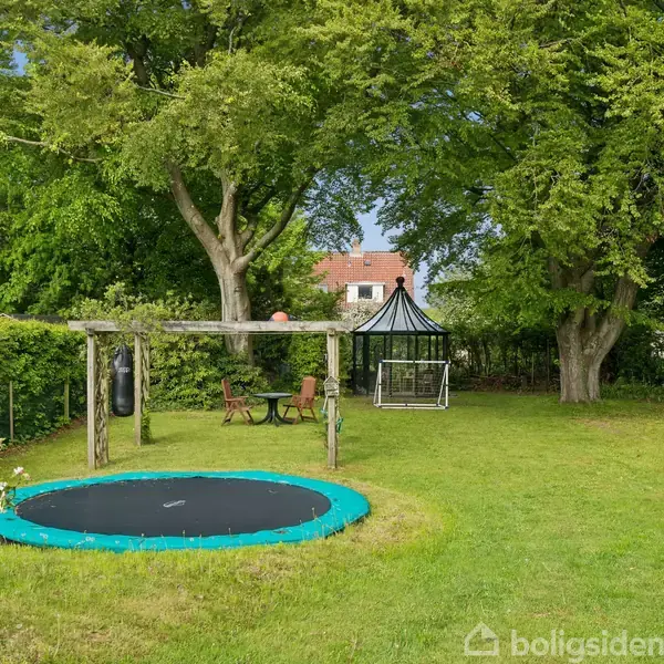 En trampolin er placeret i en frodig have med grønne træer omkring. Ved siden af står en gynge og et bord med stole. I baggrunden ses en pavillon.
