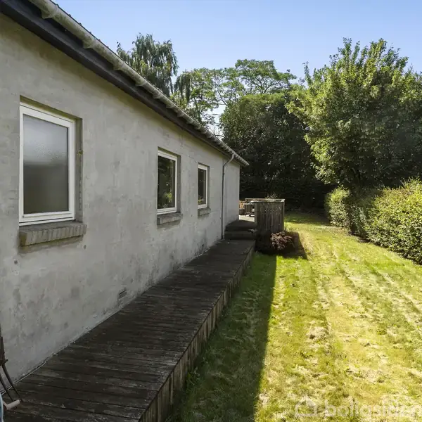 Husfacade med to vinduer; trædæk langs muren. En græsplæne strækker sig mod baghaven, omkranset af hække og træer i baggrunden. Vandkande og slange står op ad væggen.